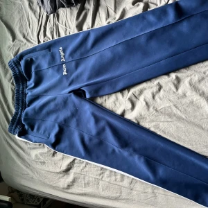 Blå Palm Angels mjukisbyxor med vita ränder - Palm Angels mjukisbyxor i marinblått med vita sidoränder och broderad logga på vänster ben. Byxorna har elastisk midja med snörning och raka ben. Tillverkade i ett glansigt syntetmaterial som ger en sportig vibe.