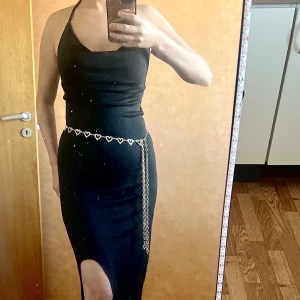 Svart långklänning med slits midjakedja  - Svart långklänning med halterneck och tight passform. Klänningen har en hög slits framtill och ärmlös design. Snyggt detaljerad med ett kedjebälte med hjärtan i midjan. Perfekt för dig som vill sticka ut med en elegant och stilren look.