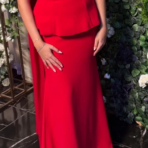 Röd långklänning med cape-detalj - Säljer en elegant röd långklänning med cape-liknande detalj över axlarna. Klänningen har en figurnära passform och en stilren, slät design utan mönster. Perfekt för dig som vill sticka ut med en klassisk och sofistikerad look.