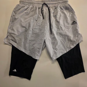 Adidas träningsshorts med tights - Snygga träningsshorts från Adidas i grått med svarta inbyggda tights. Shortsen har snörning i midjan, två fickor och Adidas-logga på både shortsen och tightsen. Perfekta för gymmet eller löprundan, tillverkade i lätt och snabbtorkande syntetmaterial.      Supersköna 