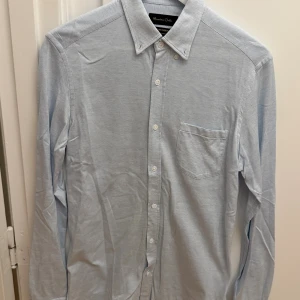 Ljusblå skjorta från Massimo Dutti M - Snygg ljusblå skjorta från Massimo Dutti i italienskt tyg. Klassisk krage med knappar, bröstficka och diskret mönster. Långärmad och tillverkad i bomull, perfekt för en clean och stilren look. Storlek M.