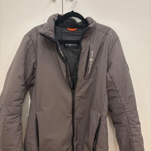 Grå vindjacka från Sail Racing - Snygg grå pufferjacka från Sail Racing med hög krage och svarta dragkedjor. Jackan har diskret logga på bröstet och coola sömdetaljer på ryggen. Perfekt för kyliga dagar och har en modern, sportig look.