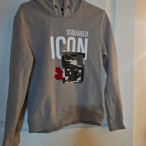 Beige hoodie från Dsquared2 med camoficka - Snygg beige hoodie från Dsquared2 med stor vit 'ICON'-text och röd lönnlöv-detalj på bröstet. Framsidan har en cool camouflagemönstrad ficka. Hoodien har långa ärmar och är tillverkad i mjuk bomull. Perfekt för dig som gillar streetstyle.