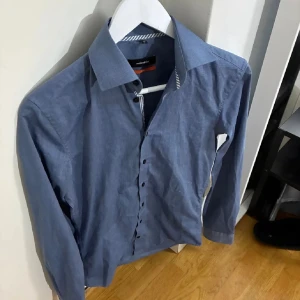 Blå skjorta från Selected Homme - Snygg blå skjorta från Selected Homme med klassisk krage och svarta knappar. Skjortan har lång ärm och är tillverkad i ett mjukt bomullstyg. Perfekt för dig som gillar stilrena och enkla plagg.