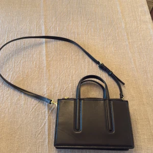 Svart axelväska i skinn med dragkedjor -  svart axelväska i skinn med justerbar rem och två handtag. Väskan har flera fack med guldfärgade dragkedjor och en boxig, strukturerad form. 