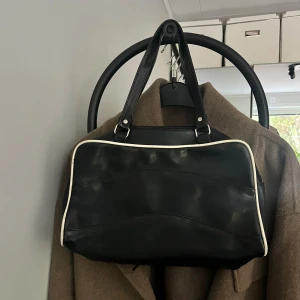 Svart handväska från Daniel Ray - Snygg svart handväska från Daniel Ray i skinnimitation med vita kanter. Väskan har dubbla handtag, dragkedja upptill och ett extra fack med dragkedja på baksidan. Insidan är fodrad med logotypmönster och har flera praktiska fack.