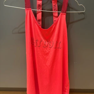 Nike röd Dri-Fit linnetopp M - Sportig röd linnetopp från Nike med Dri-Fit-material som andas. Breda axelband med Nike-logga och texten 'Just Do It' framtill. Perfekt för träning och aktiva dagar, med loose passform och snygg design.