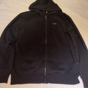 Svart hoodie med hjärtdetalj Comme des Garçons - Svart hoodie från Comme des Garçons Play med dragkedja, huva och snörning. Fram på bröstet finns det klassiska hjärtat med ögon i miniformat och en större version på ryggen. Praktiska fickor framtill och mjukt bomullsmaterial. Perfekt för en chill och stilren look.