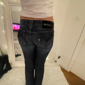 Blå jeans från BeltArt med detaljer - Säljer ett par mörkblå jeans från BeltArt med unika bakfickor med nitar och knappar. Jeansen har låg midja och raka ben, klassisk denimlook och coola detaljer som sticker ut. Perfekta för dig som vill ha något annorlunda i garderoben.