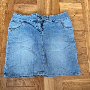 Ljusblå jeanskjol från Cue, strl 36 - Ljusblå jeanskjol från Cue med klassisk femficksdesign, dubbla knappar och snörning i midjan. Kjolen är i kort modell och har en liten slits bak. Materialet är 78% bomull, 18% polyester och 4% spandex för en bekväm passform.