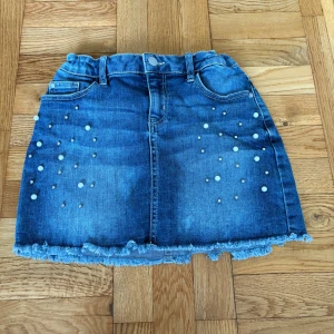 Blå jeanskjol med pärlor från Lindex - Snygg blå jeanskjol från Lindex med pärldetaljer framtill och fransad nederkant. Kjolen har klassiska jeansfickor både fram och bak samt hällor för bälte. Perfekt för dig som gillar denim med lite extra detaljer.