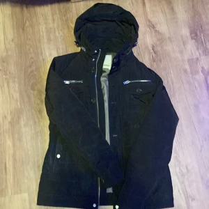 Svart vindjacka från J.Lindeberg - Svart vindjacka från J.Lindeberg med huva, dragkedja och flera fickor med både dragkedja och knappar. Jackan har en stilren look och är perfekt för blåsiga dagar. Materialet är troligtvis polyester och jackan har långa ärmar.