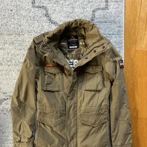 Olivgrön field jacket från Superdry - Snygg olivgrön parkajacka från Superdry med flera stora fickor, patch på ärmen och coola detaljer. Jackan har hög krage, axelklaffar och dragkedja med tryckknappar. Perfekt för dig som gillar streetstyle och utility-vibe.