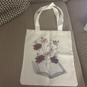 Vit tygkasse med blommor och bokmotiv - Superfin vit tygkasse med tryck av en öppen bok där blommor och fjärilar växer upp ur sidorna. Väskan har långa handtag och är perfekt att slänga över axeln. Materialet är kraftig bomull och motivet går i rosa, rött och svart.