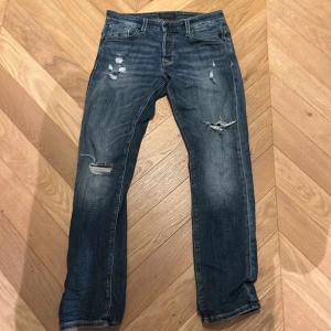 Jack and Jones jeans - Säljer ett par blå jeans med slitningar och hål på låren och knäna. Jeansen har klassisk femficksdesign, raka ben och normal passform. Materialet är denim och färgen är en tvättad blå nyans med ljusare partier. Perfekta för en avslappnad och trendig look.