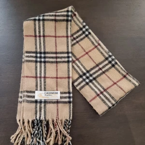 Beige rutig kashmirhalsduk - Snygg beige halsduk i 100% kashmir från Royal Rossi med klassiskt rutigt mönster i svart, vitt och rött. Mjukt material och fransar i ändarna ger en stilren och tidlös vibe. Perfekt accessoar för att lyfta din outfit under kyliga dagar.
