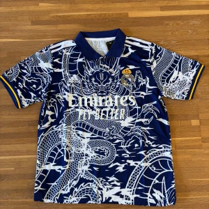 Real Madrid x Adidas blå draktröja | Lätt & ventilerande - Säljer en Real Madrid fotbollströja från Adidas i mörkblått med vit drakdesign över hela tröjan. Tröjan har klassisk krage med knappar, broderat klubbmärke och gula detaljer på ärmsluten. Tillverkad i lätt och ventilerande polyester – perfekt för match, träning eller streetwear.  ✔️ Lätt och andas ✔️ Kortärmad, regular fit ✔️ Aldrig använd  Storlek: M Skick: Ny / aldrig använd
