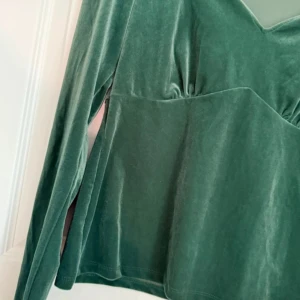 Grön sammetsblus från Vila - Mörkgrön sammetsblus från Vila med lång ärm och v-ringad hals. Toppen har en figurnära passform upptill och lätt utsvängd nertill. Mjuk och lyxig känsla i materialet, perfekt för att lyfta din outfit.