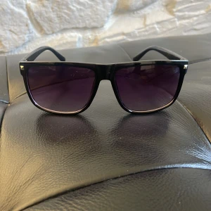 Svarta solglasögon med lila tonade glas - Snygga svarta solglasögon med rektangulär form och lila tonade glas. Klassisk och stilren design med diskreta metalldetaljer i hörnen. Perfekta för att lyfta din look under soliga dagar.