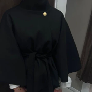Svart kappa med bälte och guldknapp - Stilren svart kappa med vid siluett och knytbälte i midjan. Jackan har hög krage och en dekorativ guldknapp vid halsen. Perfekt för dig som gillar minimalistisk och elegant look.