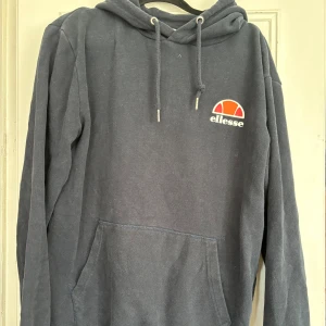 Mörkblå hoodie från Ellesse - Säljer en mörkblå hoodie från Ellesse med klassisk logga på bröstet. Tröjan har huva med snörning och en stor magficka. Perfekt för dig som gillar en sportig och avslappnad stil.