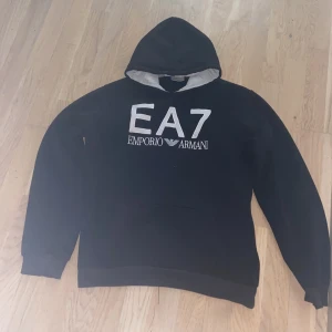 Svart EA7 Emporio Armani hoodie - Svart hoodie från EA7 Emporio Armani med stor vit logga broderad på bröstet. Klassisk huva med ljus insida och långa ärmar. Perfekt för dig som gillar stilrena och exklusiva streetwear-plagg.