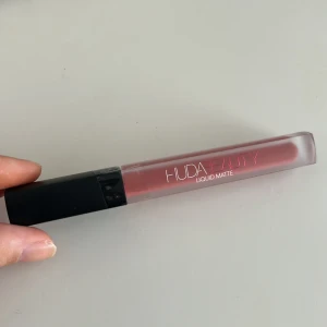 Huda Beauty Liquid Matte Bombshell - Flytande läppstift från Huda Beauty i nyansen Bombshell. Färgen är en mjuk nude-rosa med matt finish. Kommer i en stilren, fyrkantig tub med svart lock och transparent kropp som visar färgen. Perfekt för dig som gillar trendiga, matta läppar. Ordinarie 339, pris 150kr