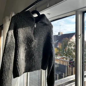 Mörkgrå stickad half zip tröja - Mysig mörkgrå stickad tröja med half zip-dragkedja och hög krage. Tröjan är ribbstickad och har en croppad passform med långa ärmar. Perfekt för kyliga dagar när du vill vara både bekväm och snygg.