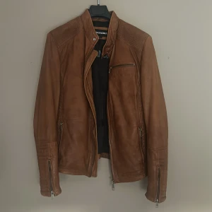 Brun skinnjacka Rockandblue M - Snygg brun skinnjacka från Rockandblue i storlek M. Jackan har coola detaljer med dragkedjor vid ärmslut och fickor, samt quiltade partier på axlar och sidor. Fodret är svart med orange kantband och flera innerfickor. Perfekt för dig som gillar stilren och edgy look.