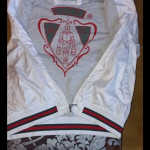 Vit vindjacka från Gucci äkta storlek lek xxl men passar L - Snygg vit vindjacka från Gucci med diskret GG-mönster i tyget, klassiska gröna och röda ränder på muddar och midja samt dragkedja framtill. Jackan har lång ärm och liten broderad logga på bröstet. Perfekt för dig som vill sticka ut med en lyxig streetstyle.