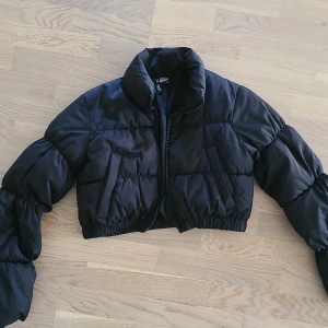 Svart croppad pufferjacka Divided XS - Svart croppad pufferjacka från Divided by H&M i storlek XS. Jackan har hög krage, dragkedja framtill och två snedställda fickor. Puffiga ärmar och elastisk nederkant ger en trendig och cool look. Perfekt för kyliga dagar när du vill sticka ut.