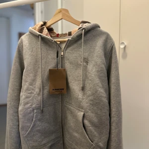 Burberry Check Zip Hoodie - HypeTorget Grå hoodiejacka från Burberry med klassiskt rutigt foder i beige, svart och rött. Jackan har dragkedja, snörning i huvan och broderad Burberry-logga på bröstet. Tillverkad i mjuk bomull- och polyesterblandning, perfekt för en chill men stilren look. Storlek M, passar även Small för en något oversized fit.  🖤 Notera: Produkten postas endast – ingen mötesaffär. 📌 Läs gärna bio på min Plick-profil innan köp. 