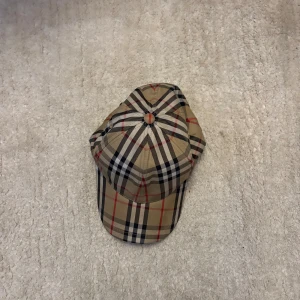 Burberry rutig beige keps - Snygg keps från Burberry med klassiskt rutigt mönster i beige, svart, vitt och röda linjer. Kepsen har böjd skärm och justerbar passform. Perfekt för dig som vill ha en ikonisk accessoar med lyxig känsla och street vibe.