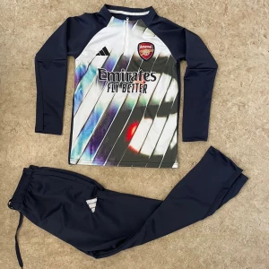 Arsenal långärmad fotbollströja Adidas - Snygg Arsenal fotbollströja från Adidas med lång ärm och zip framtill. Framsidan har ett coolt randigt mönster i vitt, svart, blått och rött med klubbmärke och Emirates Fly Better-tryck. Materialet är lätt och stretchigt, perfekt för träning eller match.