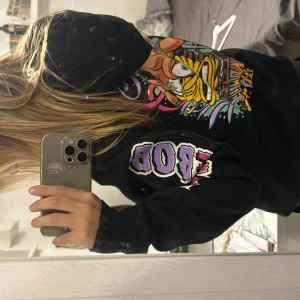 Svart Spongebob hoodie med tryck - Svart hoodie med färgglatt Spongebob och Patrick-tryck på ryggen och mindre tryck på bröstet. Texten 'SPONGEBOB' i lila och rött på båda ärmarna. Klassisk huva med snörning och känguruficka framtill. Perfekt statement-plagg för dig som gillar tecknade serier.