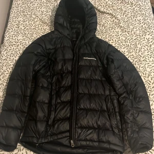 Svart dunjacka Peak Performance S - Svart dunjacka från Peak Performance i storlek S. Jackan har quiltad design, huva och dragkedja framtill. Klassisk logga på bröstet och ärmen. Perfekt för kalla dagar och riktigt snygg till streetwear-stilen.