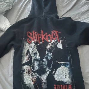 Svart Slipknot hoodie med tryck - Svart hoodie med Slipknot-tryck både fram och bak. Framsidan har rött Slipknot-logo och texten '2001 IOWA 2016' med grafiskt mönster. Baksidan har stort tryck med bandets logga och motiv från albumet Iowa. Klassisk känguruficka och huva.