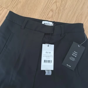 Svarta kostymbyxor med slits från NA-KD, strl 34 - Svarta kostymbyxor från NA-KD i rak modell med hög midja. Byxorna har dragkedjor vid bensluten för en snygg detalj och är tillverkade i ett slätt, återvunnet material. Perfekta för en clean och stilren look.
