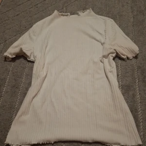 Vit ribbad topp från ONLY - Säljer en vit ribbad topp från ONLY i storlek M. Toppen har korta ärmar, lätt vågiga kanter och en smal passform. Perfekt att styla med jeans eller kjol för en clean look.