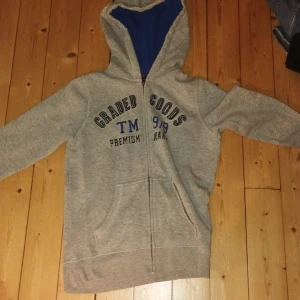 Grå hoodie från Pepperts med tryck - Grå zip-hoodie från Pepperts med blått innerfoder i huvan och tryck på bröstet. Tröjan har dragkedja, känguruficka och långa ärmar. Perfekt för chill dagar eller när du vill ha en enkel och snygg stil.