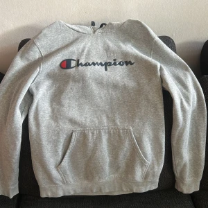 Grå hoodie från Champion - Säljer en grå hoodie från Champion med klassisk logga framtill. Tröjan har huva med snörning och en stor magficka. Perfekt för dig som gillar en enkel och sportig stil. Långärmad och i mjukt bomullsmaterial.