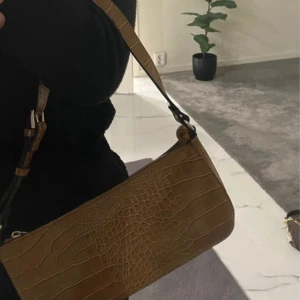 Brun axelväska i krokopräglat skinn - Säljer en brun axelväska med croco-mönster och stilren form. Väskan har justerbar rem och stängs med dragkedja upptill. Perfekt storlek för det viktigaste och ger en trendig touch till din outfit.