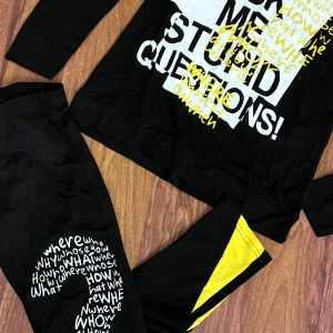 Svart långärmad pyjamas med tryck - Svart långärmad pyjamas med stort grafiskt tryck i vitt och gult framtill med texten 'DON'T ASK ME STUPID QUESTIONS!' och ord i olika stilar. Tröjan har gula detaljer vid axlarna och är i mjukt material, perfekt för en avslappnad och cool look.