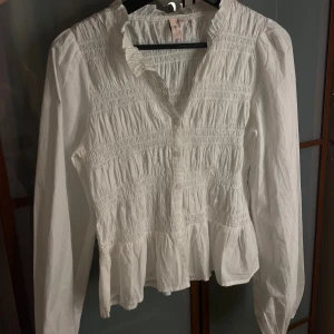 Vit volangskjorta med smock - Supersöt vit skjorta från NELLY med smockad överdel, volangkant nertill och lätt rynkade axlar. Skjortan har långa ärmar, knappar framtill med liten volangkrage. Perfekt för dig som gillar romantisk stil.