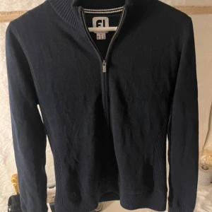 Mörkblå kofta från FootJoy med dragkedja - Snygg mörkblå kofta från FootJoy med hel dragkedja framtill och ribbade detaljer vid krage, sidor och ärmslut. Klassisk och stilren design, perfekt för lager-på-lager. Långärmad och i mjukt material som känns skönt mot huden.