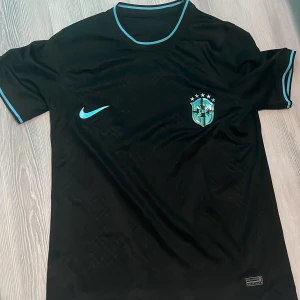 Svart Brasilien fotbollströja Nike - Snygg svart Brasilien fotbollströja från Nike med turkosa detaljer och emblem. Tröjan har korta ärmar, rund hals och är tillverkad i ett lätt, ventilerande material. Perfekt för dig som vill sticka ut på planen eller på läktaren.