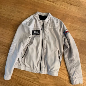 Porsche Design Martini Racing jacka M - SUPERSNYGG ALDRIG ANDVÖND FAM JACKA KAN ÄVEN ANDVÄNDAS I REGN HELT NY INGA FLÄCKAR SPRUKNA SÖMMAR OSV JÄTTE SKÄN OCH FIN PASSAR TILL ALLT OCH ALLA!! Snygg ljusgrå vindjacka från Porsche Design med Martini Racing-detaljer. Jackan har broderade patches, dragkedja framtill och quiltade partier på axlar och armbågar. Materialet är lätt och syntetiskt, perfekt för dig som gillar racingstil och sportiga vibbar.