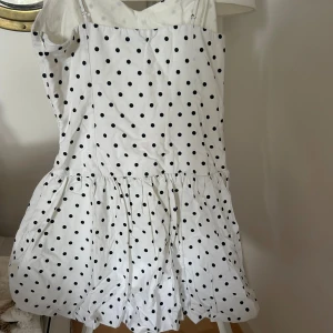 Vit prickig kortklänning med axelband - Supersöt vit kortklänning med svarta prickar och tunna axelband. Klänningen har en voluminös kjol med rynk och en feminin v-ringad överdel. Perfekt för dig som gillar lekfulla mönster och en lätt, somrig look.