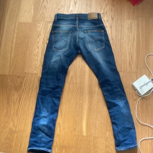 Nudie Jeans blå jeans, storlek 29/30 - Säljer ett par klassiska blå jeans från Nudie Jeans med snygga slitningar och orangea sömmar. Modellen har raka ben och fem fickor, samt Nudie-logga på bakfickan. Jeansen är i stretchigt bomullsmaterial och har normal passform.