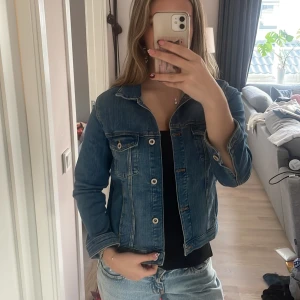 Blå jeansjacka från Zara - Snygg klassisk blå jeansjacka från Zara med raka linjer, bröstfickor med knappar och krage. Jackan har långa ärmar och silverfärgade knappar framtill. Perfekt att slänga över en hoodie eller t-shirt för en chill look.